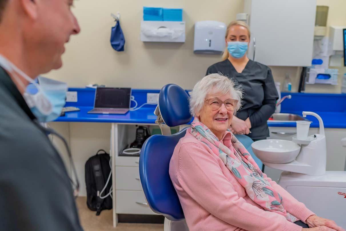 Smiling retired dental patient sitting in chair with expert dentists in background and foreground.