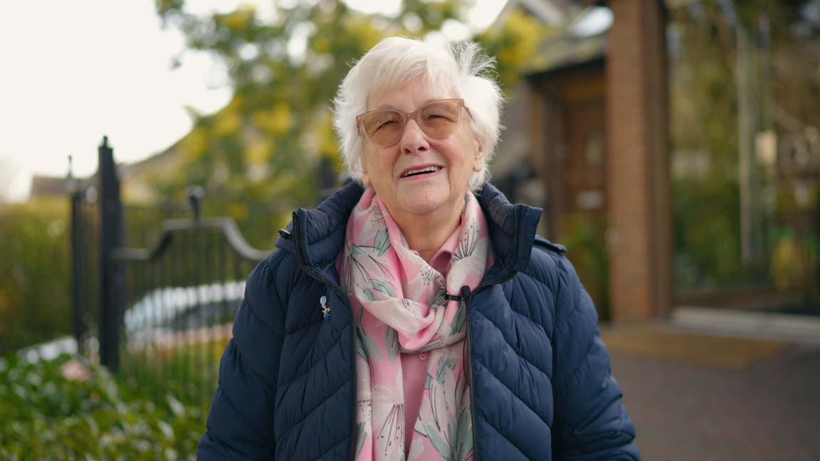 Smiling older woman outdoors after dental implant treatment, wearing a navy coat and pink scarf, showing restored confidence and comfort.
