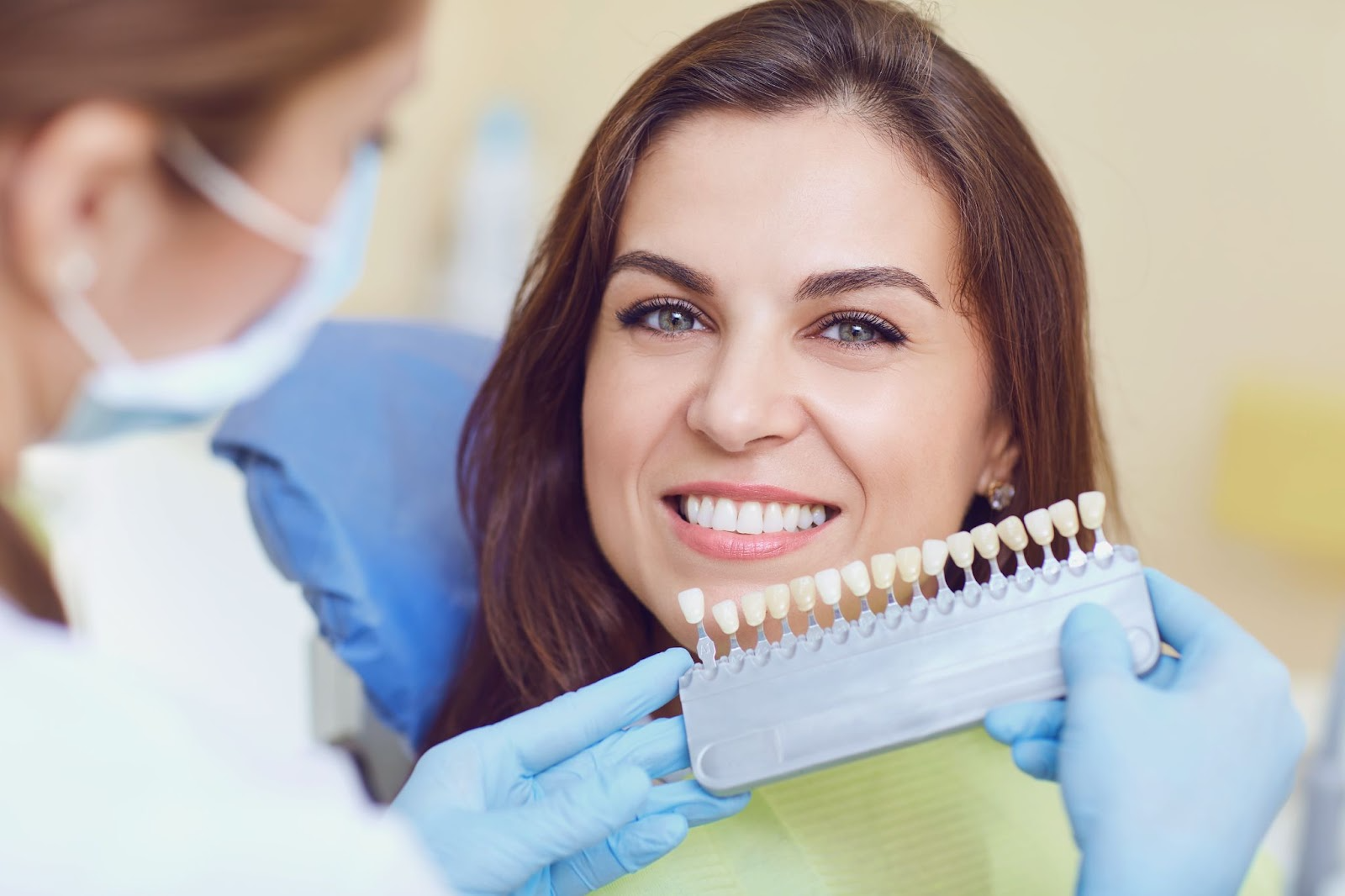 Female patient smiling as dentist compares tooth shades for teeth whitening treatment using a shade guide.