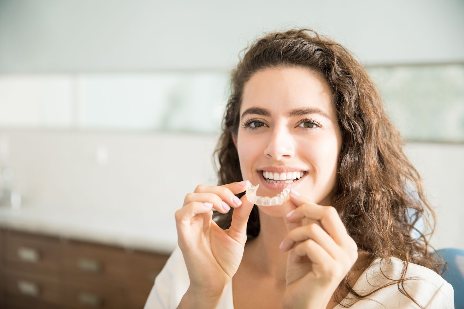 Woman holding a clear Invisalign aligner, smiling confidently in a dental clinic setting.